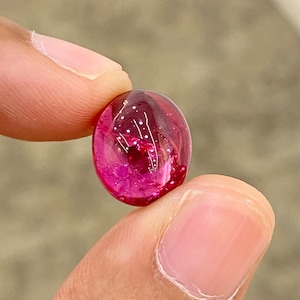 May include: A vibrant pink oval-shaped gemstone with a smooth, polished surface. The gemstone is translucent and reflects light, creating a sparkling effect.