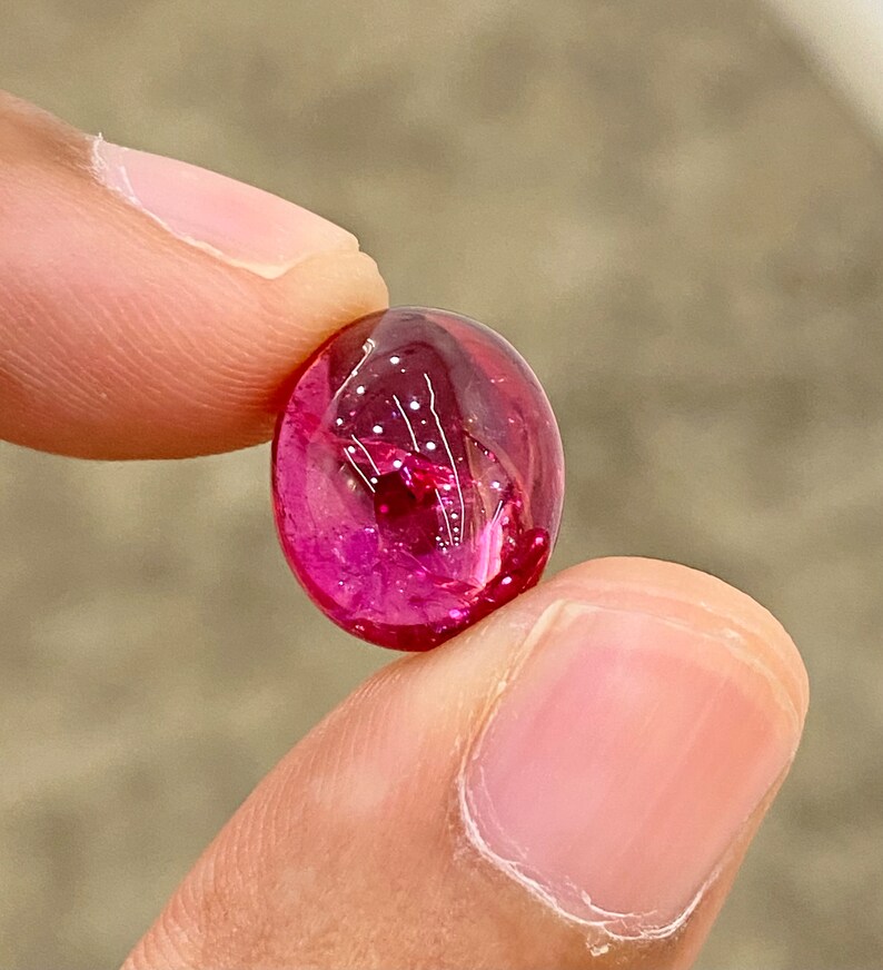 May include: A vibrant pink oval-shaped gemstone with a smooth, polished surface. The gemstone is translucent and reflects light, creating a sparkling effect.