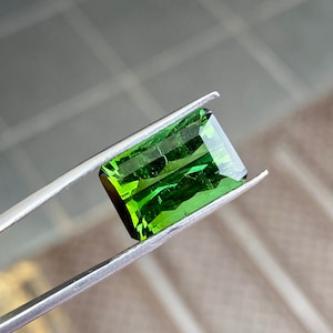 May include: A vibrant green gemstone with a rectangular cut, held by tweezers. The gemstone has a slightly cloudy appearance, with visible inclusions.