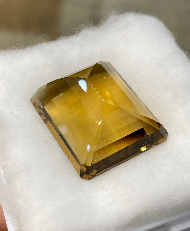 May include: A large, rectangular, faceted gemstone with a golden brown color. The gemstone is cut in a step-cut style and has a high polish.
