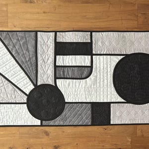 May include: A rectangular quilted table runner with a geometric design in shades of gray, black, and white. The runner features three large black circles and various rectangular and curved shapes. The design is outlined in black.