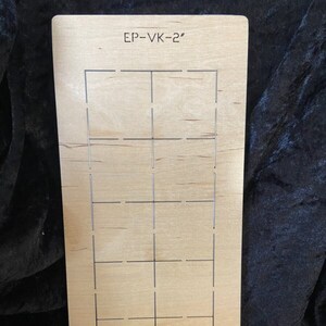 May include: A rectangular wooden die cut with a grid pattern and the text "EP-VK-2". The die cut is made of light-colored wood and has a series of cut lines forming a grid of rectangular shapes. The die cut is approximately 2 inches in length.