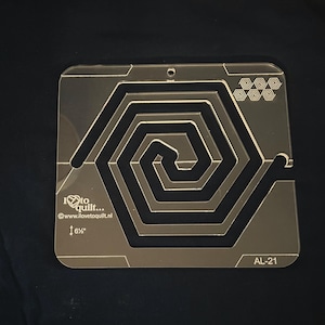 Pode incluir: Um modelo de acolchoado de plástico transparente com um design em espiral. O modelo tem 16,5 cm de largura e tem o texto "I love to quilt... www.ilovetoquilt.nl" e "AL-21" impresso nele.