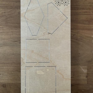 Peut inclure: Gabarit de découpe en bois avec le texte "Fabric Scarlet EP-8P-SC-FB-2" et plusieurs formes géométriques découpées. Les formes comprennent un hexagone, un losange, un carré et un motif floral décoratif. Le gabarit est en bois clair.