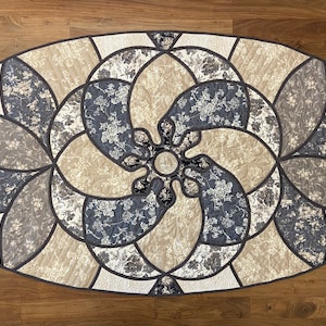 Puede incluir: Camino de mesa acolchado de forma ovalada con un estampado floral en tonos beige, gris y azul marino. El diseño presenta un motivo floral central y está bordeado por un ribete gris oscuro. El camino de mesa está sobre una superficie de madera.