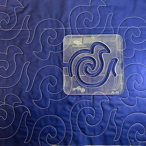 Pode incluir: Uma imagem em close-up de um tecido acolchoado azul com um padrão de redemoinhos brancos repetidos. Uma régua de quilting de acrílico transparente com um design semelhante a um pássaro é colocada sobre o tecido. A régua tem o texto "18" e "AL-23".