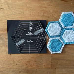 Op de afbeelding: Een doorzichtige, zeshoekige liniaal van plastic met markeringen voor 1/2, 2 1/2, 3, 3 1/2 en 4 inch. De liniaal ligt op een zwart oppervlak. Een zeshoekig patchwork van stof ligt op een houten oppervlak.
