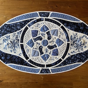 May include: Oval quilted table runner in shades of blue and white. The center features a floral design, surrounded by a geometric pattern. The runner is approximately 30 inches long and 18 inches wide.