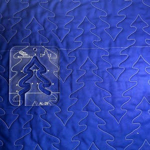 May include: A blue quilted fabric with a repeating tree pattern stitched in white thread. A clear plastic quilting template with a tree design is placed on the fabric. The template is marked with measurements.