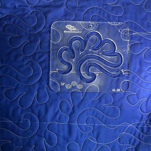 Könnte beinhalten: Nahaufnahme eines blauen, gesteppten Stoffes mit einem Wirbelmuster. Eine transparente Acryl-Quiltschablone liegt obenauf und weist ein dekoratives Design auf. Die Schablone ist mit dem Text "I love to quilt..." und "AL-26" versehen.