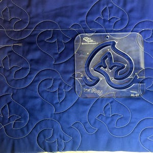 May include: A close-up of a blue quilted fabric with a repeating floral pattern stitched in white thread. A clear acrylic quilting template, 17.5 inches, is placed on the fabric, showing the design.