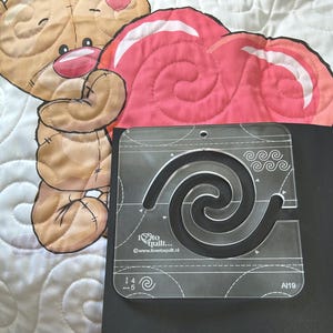 May include: A clear plastic quilting template featuring a spiral design. The template is labelled "I love to quilt..." and includes the website address "www.ilovetoquilt.nl". The template also has a scale marked "1/4 - 1/5" and the number "A19".