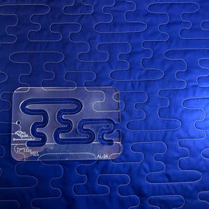 Könnte beinhalten: Nahaufnahme eines blauen Quilts mit einem sich wiederholenden weißen Nahtmuster. Eine transparente Acryl-Quiltschablone mit geschwungenem Design liegt auf dem Stoff. Die Schablone trägt den Text "www.lovetoquilt.nl" und "AL-24".