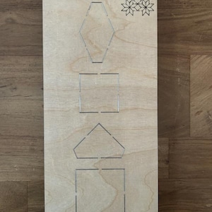 Könnte beinhalten: Eine Holzstanzschablone mit verschiedenen geometrischen Formen, darunter ein Sechseck, ein Quadrat und ein Fünfeck. Die Schablone zeigt auch ein Schneeflockenmuster und den Text "Fabric Scarlet EP-8P-SC-FB-1¼".