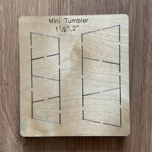 May include: A wooden craft template for a mini tumbler, featuring etched lines for cutting and shaping. The text "Mini Tumbler" is printed at the top, with size specifications of 1 1/2" and 2". The template is rectangular and sits on a wooden surface.