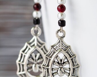 May include: Silver spider web earrings with a dangling spider charm. The earrings are made with black, white, and red beads.