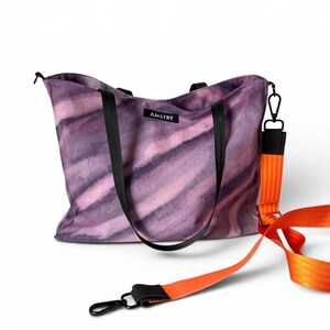 Könnte beinhalten: Eine violette und dunkelviolette Batik-Tragetasche mit schwarzen Griffen und einem schwarzen Etikett mit der Aufschrift AMETRY. Die Tasche verfügt über einen abnehmbaren, leuchtend orangefarbenen Nylon-Schultergurt mit schwarzen Metallkarabinerhaken.