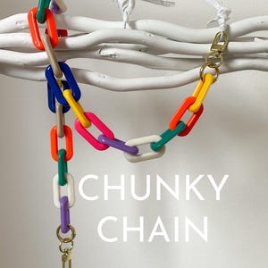 Könnte beinhalten: Eine farbenfrohe, dicke Kette aus Plastikgliedern in verschiedenen Farben, darunter Grün, Blau, Orange, Pink, Gelb und Weiß. Die Kette hängt an einer weißen Holzoberfläche und hat an beiden Enden goldene Metallverschlüsse. Der Text "CHUNKY CHAIN" ist in Weiß unter der Kette gedruckt.