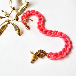 Könnte beinhalten: Ein pinkfarbenes Plastikkettenarmband mit einem goldenen Stierkopf-Anhänger. Das Armband besteht aus dicken, massiven Gliedern.