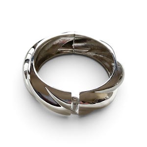 Könnte beinhalten: Ein polierter, silberfarbener Metall-Klapparmreif mit einem dicken, gedrehten oder geknoteten Design. Das Armband hat eine glatte, reflektierende Oberfläche und einen sicheren Federklappverschluss, der ein einfaches Öffnen und Schließen ermöglicht.