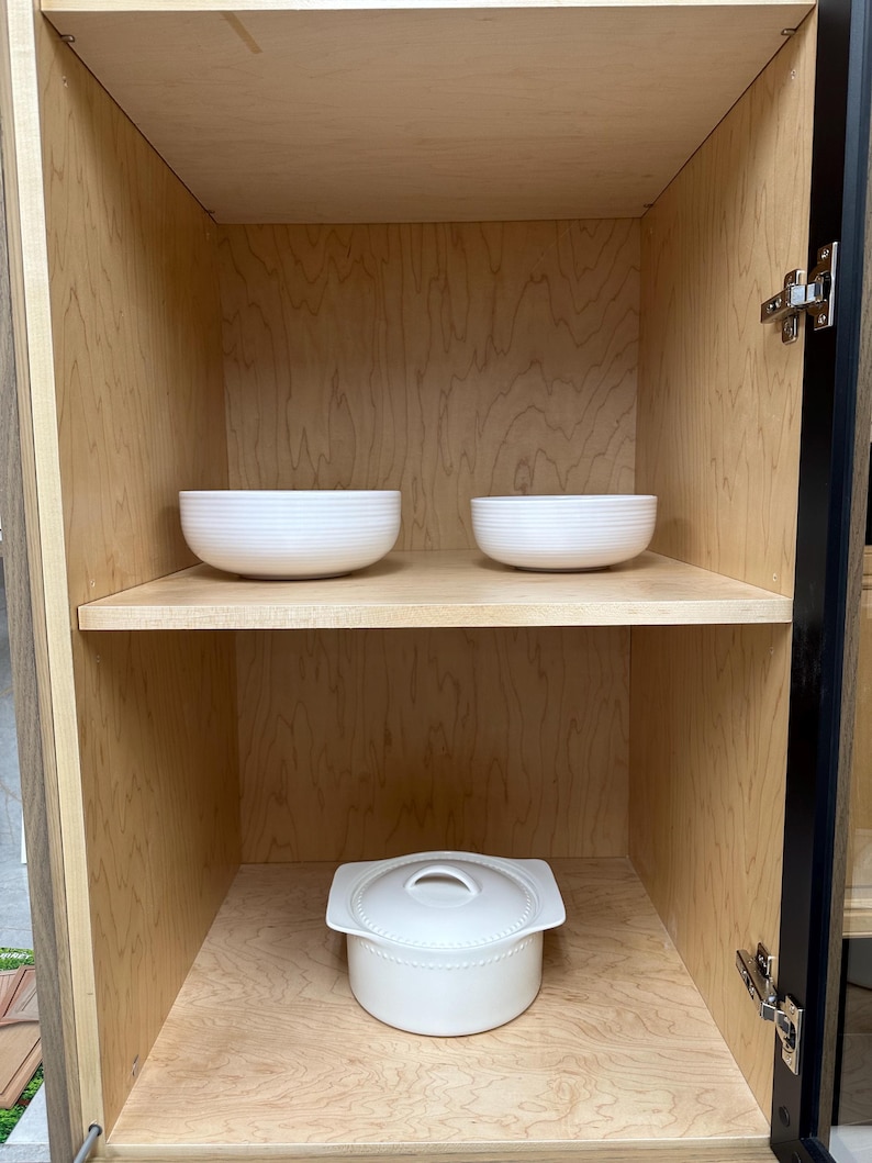 Op de afbeelding: Twee witte keramische kommen met horizontale ribbelstructuren staan op de bovenste plank van een lichtgekleurde houten kast. Daaronder, op de onderste plank, staat een witte keramische ovenschaal met deksel en zijhandgrepen.
