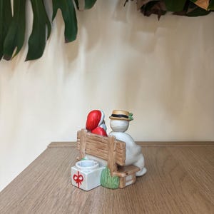 Könnte beinhalten: Eine Keramik-Weihnachtsfigur, die den Weihnachtsmann und einen Schneemann auf einer Holzbank zeigt. Der Weihnachtsmann trägt einen roten Anzug und Hut, der Schneemann einen Hut mit einem grünen Zweig. Eine Geschenkbox mit roter Schleife steht daneben.