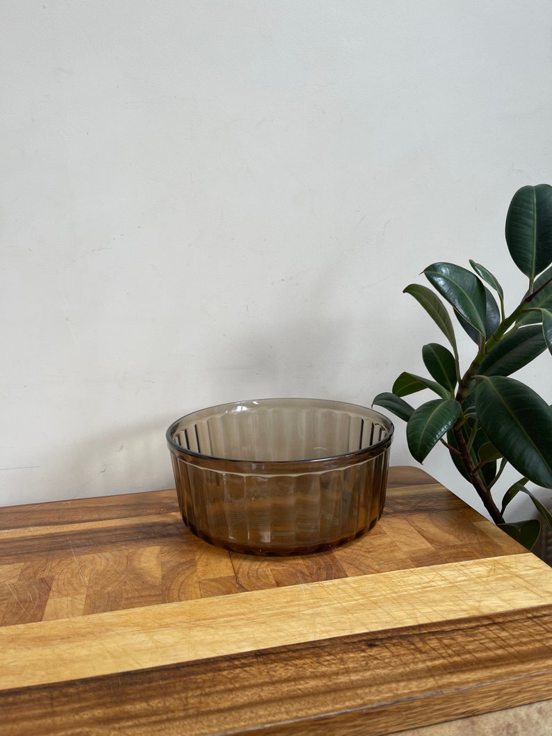 May include: A brown glass ramekin with a fluted design. The ramekin is sitting on a wooden cutting board.