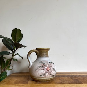 Könnte beinhalten: Eine weiße Keramikkanne mit brauner Glasur und einem Blumendesign. Die Kanne hat einen Henkel und einen runden Körper.