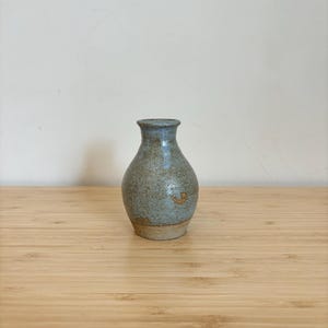 Könnte beinhalten: Eine kleine, hellblaue Keramikvase mit einem gesprenkelten Überzug. Die Vase hat einen runden Körper und einen schmalen Hals.