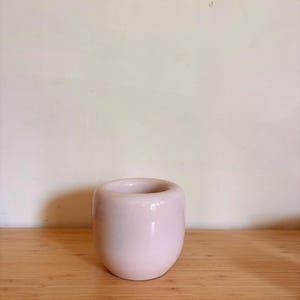 Könnte beinhalten: Eine kleine, hellrosa Keramikvase mit rundem Körper und breiter Öffnung. Die Vase hat eine glatte, glänzende Oberfläche und steht auf einer hellbraunen Holzoberfläche. Der Hintergrund ist eine schlichte, cremefarbene Wand.