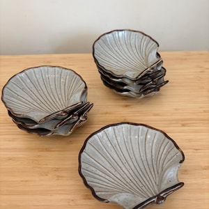 Peut inclure: Un ensemble de six petits plats en céramique blanche en forme de coquillages avec des bords marrons. Les plats sont empilés en deux piles sur une surface en bois.