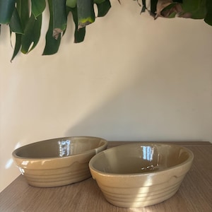 Op de afbeelding: Twee ovale, beige keramische kommen met een geribbeld ontwerp. De kommen staan op een houten oppervlak, met een plant op de achtergrond. De kommen hebben een glanzende afwerking en zijn leeg.