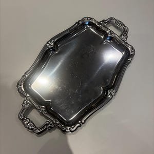 Könnte beinhalten: Ein rechteckiges Serviertablett aus Silber mit aufwendigen Details und zwei Griffen. Das Tablett hat eine reflektierende Oberfläche und einen dekorativen Rand. Die Griffe sind ebenfalls mit einem Rollendesign versehen.