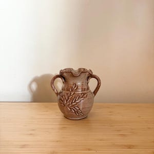 Könnte beinhalten: Eine kleine, braune Keramikvase mit zwei Henkeln und einem gewellten Rand. Die Vase hat ein Design aus braunen Blättern auf der Vorderseite.