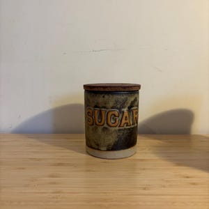 Könnte beinhalten: Eine Zuckerdose aus Keramik mit einem Holzdeckel. Die Dose ist grün und braun mit dem Wort "SUGAR" in orangefarbenen Buchstaben.