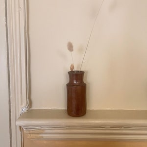 Può includere: Un vaso in ceramica marrone con collo stretto e base larga, che contiene tre steli di erba della pampa secca.