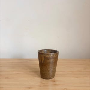 Könnte beinhalten: Eine kleine, braune Keramiktasse mit strukturierter Oberfläche. Die Tasse hat eine leicht konische Form und steht auf einer hellen Holzoberfläche. Der Hintergrund ist eine schlichte, cremefarbene Wand.