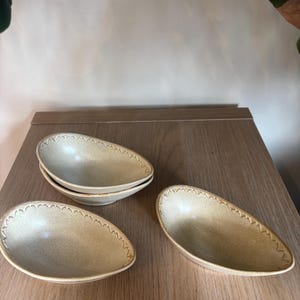 Op de afbeelding: Vier beige, ovaalvormige keramische kommen met een getextureerd oppervlak en een decoratief zigzagpatroon langs de rand. De kommen zijn gerangschikt op een houten oppervlak, wat hun unieke ontwerp benadrukt.