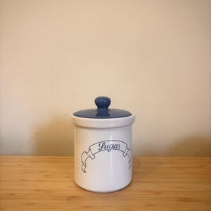 Könnte beinhalten: Ein weißer Keramik-Zuckertopf mit blauem Deckel und Knauf. Der Topf hat ein blaues Banner-Design mit dem Wort "Sugar" in Schreibschrift. Der zylindrische Topf steht auf einer Holzoberfläche.