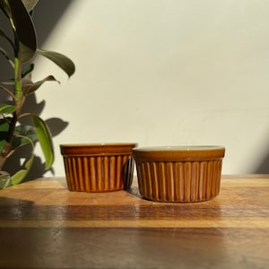 Könnte beinhalten: Zwei braune Keramik-Ramekins mit geripptem Design. Die Ramekins stehen auf einem Holzbrett.