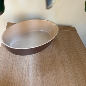Puede incluir: Una fuente de horno ovalada de cerámica marrón y beige. La fuente tiene un interior beige claro y un exterior marrón más oscuro. La fuente está sobre una superficie de madera.