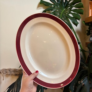Op de afbeelding: Ovale serveerschaal met een crèmekleurig midden en een dieprode rand. De schaal wordt door een hand vastgehouden. Op de achtergrond een grote groene plant.