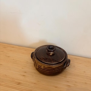 Könnte beinhalten: Ein brauner Keramiktopf mit Deckel und zwei Griffen. Der Topf hat ein gestreiftes Muster und steht auf einer Holzoberfläche.