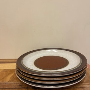 Może przedstawiać: Stos czterech ceramicznych talerzy z brązowo-białym wzorem. Talerze mają brązową obwódkę, biały środek i ciemnobrązowy okrąg pośrodku. Talerze są ułożone na drewnianej powierzchni.