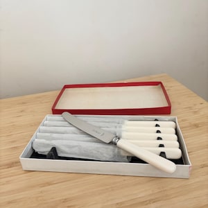 Könnte beinhalten: Ein Set mit sechs Buttermessern mit weißen Griffen in einer weißen Box mit rotem Deckel. Die Messer sind in Seidenpapier eingewickelt und die Box steht auf einer Holzoberfläche.
