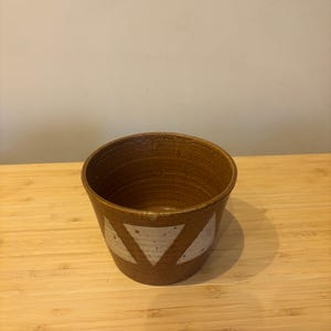 Könnte beinhalten: Ein brauner Keramiktopf mit strukturierter Oberfläche und weißen Dreiecksdesigns. Der Topf ist zylindrisch und steht auf einer Holzoberfläche. Der Topf wird wahrscheinlich für Pflanzen oder zur Aufbewahrung verwendet.