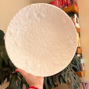 Könnte beinhalten: Große, cremefarbene Keramik-Servierschüssel mit geprägtem Blatt- und Eicheldesign. Die runde Schüssel hat eine breite, flache Form, die sich zum Servieren von Speisen oder als dekoratives Element eignet. Die Schüssel wird von einer Person gehalten.