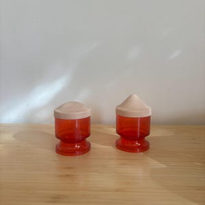 Könnte beinhalten: Zwei Vintage Salz- und Pfefferstreuer. Die Streuer bestehen aus transparentem rotem Kunststoff mit einem beigefarbenen Deckel. Sie haben ein klassisches Design mit rundem Sockel und zylindrischem Körper. Einer hat eine konische Spitze.