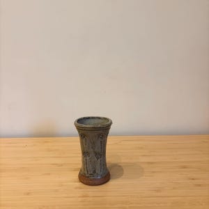 Könnte beinhalten: Eine handgefertigte Keramikvase mit zylindrischer Form und ausgestelltem Oberteil. Die Vase ist überwiegend graugrün mit einem braunen Sockel und weist ein Blumendesign auf. Die Vase ist etwa 13 cm hoch.