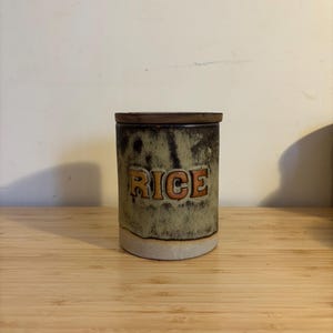 Könnte beinhalten: Eine Keramikdose mit einem Holzdeckel, in Grün- und Brauntönen bemalt. Die Dose trägt die Aufschrift "RICE" in orangefarbenen Buchstaben.